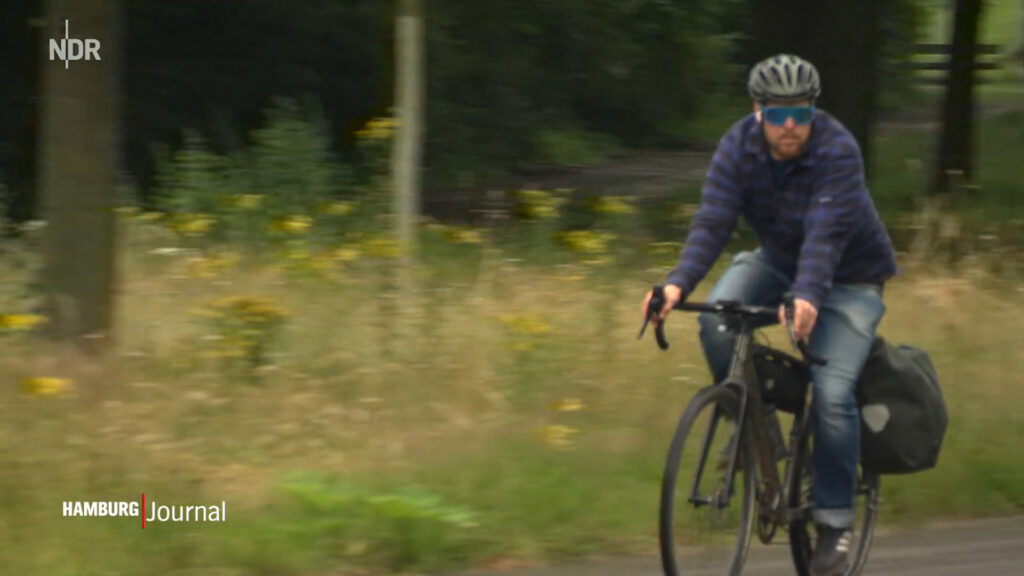 Männliche Person auf einem Gravelbike, im Hintergrund durch Motion Blur verwischtes Wegesgrün und Bäume. Als Overlay in der linken oberen Ecke das NDR-Logo, in der linken unteren das Hamburg-Journal-Logo.